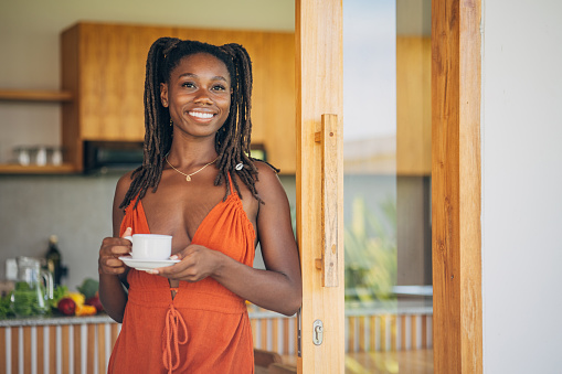 Black woman enjoys a moment of peace with a cup of coffee in her cozy, sunlit kitchen. She wears an orange dress and holds a white cup and saucer. The wooden cabinets and countertops in the background suggest a warm, inviting space. Natural light floods the room through an open door or window.
