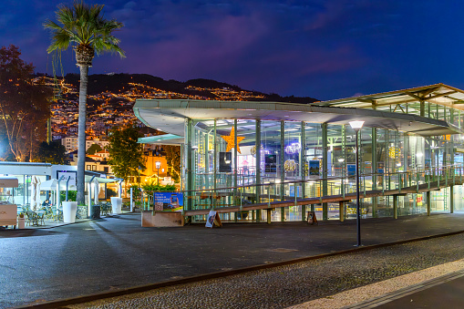 Evening view of the illuminated lower Cable Car gondola station with the city lights of Monte visible in the distance along the coast Funchal, Portugal, on the island of Madeira, Canary Islands. The base station is located in the garden of Almirante Reis, in the historic area of Funchal, with the top, in Monte, halfway between Largo das Babosas and Jardins do Monte.