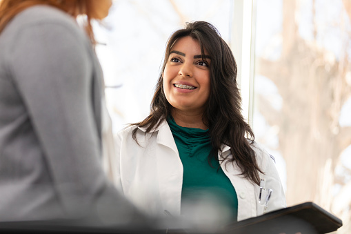 The focus of this low angle view is on the young adult female doctor smiling as she listens carefully to the unrecognizable female patient.