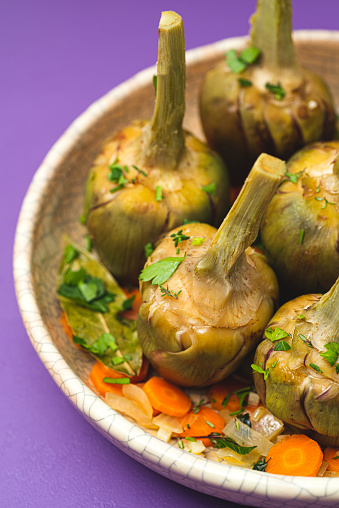 French artichokes à la Barigoule or Carciofi alla provenzal. Provencal artichokes stewed with carrots, onions, white wine, bay leaves and Provençal herbs. Spring recipes from southern France. Lavender background, close up