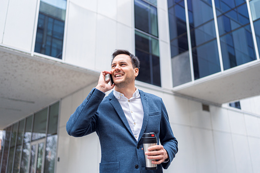 Front view of an young Caucasian businessman talking on mobile phone and holding thermos with a coffee, in front of the corporate building