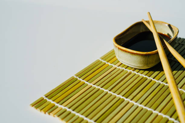 In the image, there are a pair of chopsticks resting on a bamboo sushi rolling mat, next to a small bowl filled with soy sauce. The natural textures of the bamboo and the traditional utensils evoke the essence of Japanese cuisine and the art of sushi making.