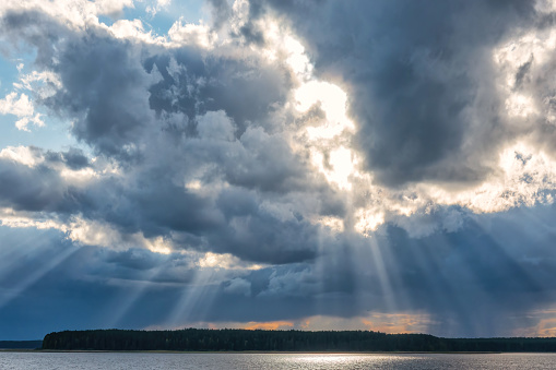 The sun's rays break through the dark clouds over the lake. A strip of forest is visible on the shore. Scenery.