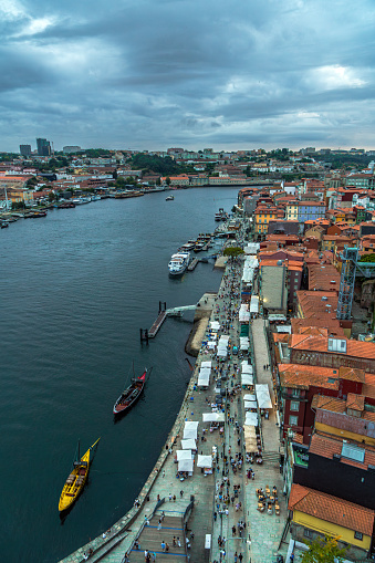 Aerial drone view of the Douro river and the Porto seafront with the old houses, and classic boats at the dock; restaurants and tourists strolling along the river walk at sunset under a cloudy sky.