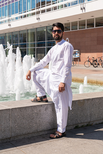 A Pakistani man with a dark beard in national white clothes stands near a fountain