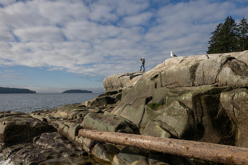 Distant view of woman standing on rock outcrop and taking photo of ocean