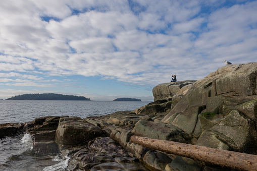 Distant view of woman standing on rock outcrop and taking photo of ocean