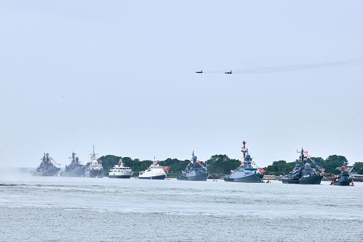 Military fighter jets are flying over Russian naval forces parade warships along coastline, seafaring tradition of military ships formation at Navy Day, nautical spectacle of russian sea power