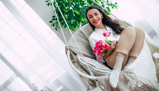 Beautiful smiling tender brunette woman with bunch of flowers is relaxing and swinging on hanging chair. Present in womens day from beloved man. St. Valentines day