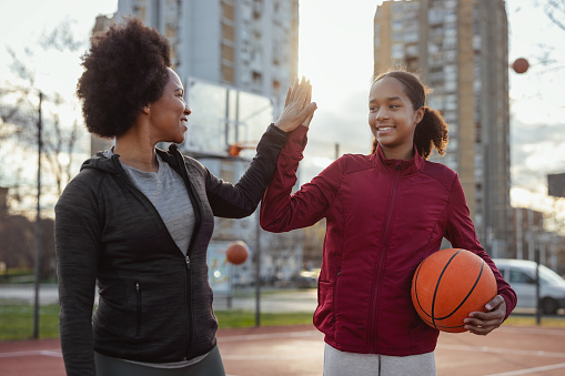 Portrait of smiling african american teenage daughter giving high five to her mother while playing basketball at the playground. Sport, people and healthy lifestyle concept