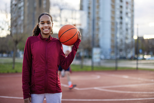 Confident african american teenage female athlete in sportswear holding basketball looking at camera on an outdoor basketball court