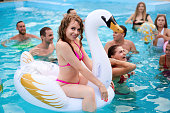 Pretty woman in bikini swimsuit hanging out on inflatable white swan mattress on night pool party. Friends partying with cocktails in holiday villa.