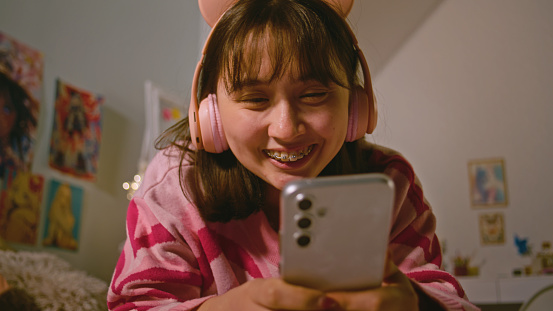 Teenage girl in headphones talks by video call and chats using mobile phone while lying on the bed in cozy bedroom. Mongolian girl spending leisure time and having fun at home. Lifestyle concept.