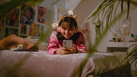 Asian teen girl in headphones talks by video chat, surfs the Internet using mobile phone while lying on the bed in cozy bedroom. Girl spending leisure time and having fun at home. Lifestyle concept.