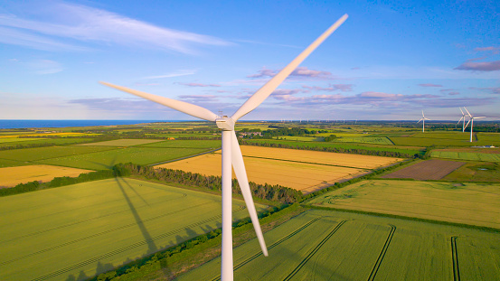 AERIAL, CLOSE UP: Rotating wind turbine on a wind farm in the English countryside. Spinning white blades of a windmill above picturesque farmland. Harnessing the wind to generate electric energy.