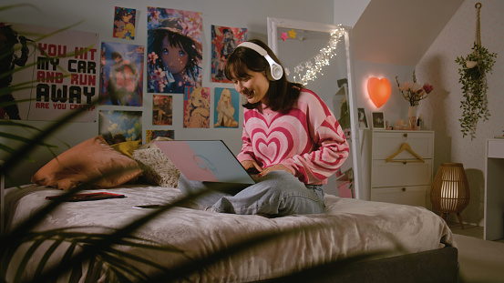Asian teenage girl in headphones listens to music, chats and surfs the Internet on laptop while sitting on the bed in her bedroom. Girl spending leisure time and having fun at home. Lifestyle concept.