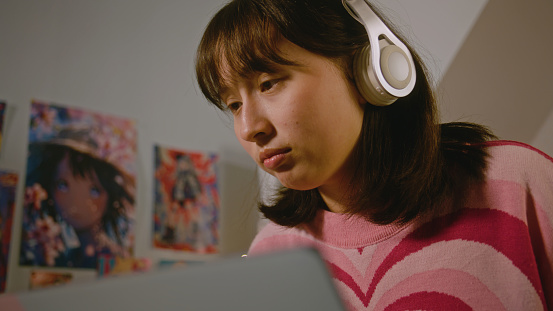 Mongolian teenage girl in headphones talks by video call on laptop while having online lesson sitting in her cozy bedroom. Student girl doing remote learning at home. Lifestyle and distance education.