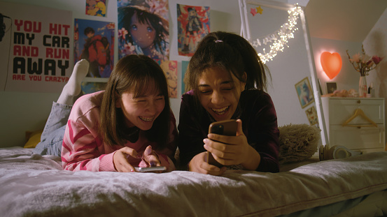 Two multicultural besties in the bedroom laugh and talk while watching funny videos using mobile phone. Joyful teenage girls spend leisure time at home lying on bed. Best friends having fun together.