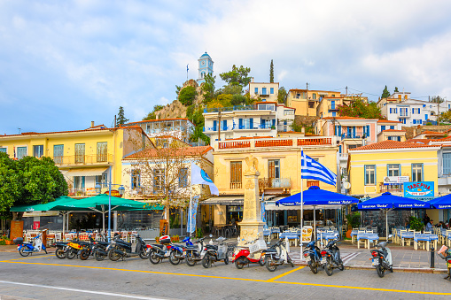 Sidewalk cafes, shops and restaurants with the blue clock tower on top of the hill behind, at the colorful seaside Greek village of Poros, on the Saronic island of Poros, Greece.