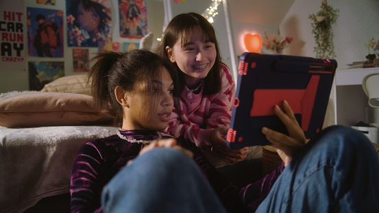 African American girl sits at floor, surfs internet using tablet. Mongolian teen lies on bed, watches content with friend. Multi ethnic girls together spend leisure time at home. Friends relationship.
