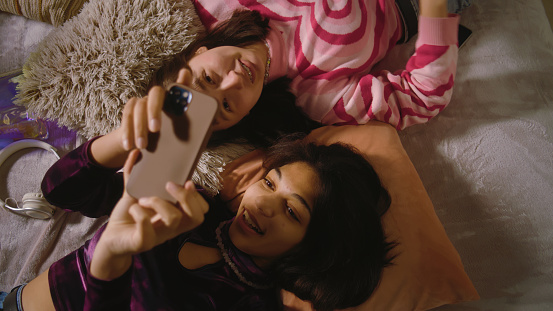 Two multiethnic best friends in the bedroom laugh and scroll through social media using mobile phone. Happy teenage girls spend leisure time at home lying on the bed. Besties having fun together.