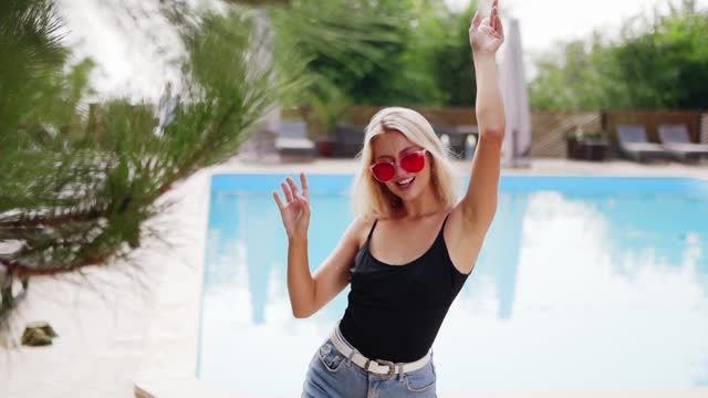 Blonde hair woman dancing for photoshoot in red sunglasses, shorts and swimwear at swimming pool in resort beach club with sun loungers and pine-trees. Model posing and flirting on holidays.