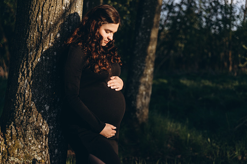 A pregnant woman is shown leaning against a tree trunk in a forest setting.