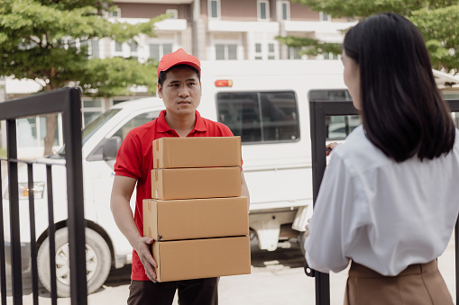 Young Asian woman receives a cardboard box from the postman at home who is smiling and happy. The courier delivers the item urgently. Online store, paper containers, postman, delivery service, parcels