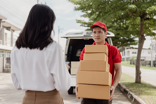 Young Asian woman receives a cardboard box from the postman at home who is smiling and happy. The courier delivers the item urgently. Online store, paper containers, postman, delivery service, parcels