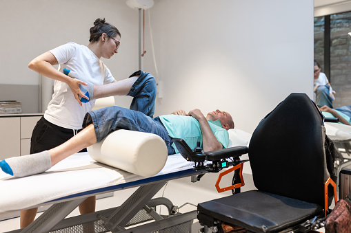 A physiotherapist examines a patient in a wheelchair. A physiotherapist does rehabilitation exercises with a disabled person and a therapy table.