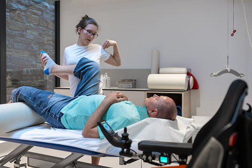 A physiotherapist examines a patient in a wheelchair. A physiotherapist does rehabilitation exercises with a disabled person and a therapy table.