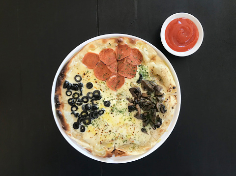 Classic baked italian pizza with pepperoni, mushrooms, mozarella cheese, and black olives served on dark black wooden rustic table.