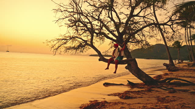 Handheld Shot of Male Tourist Sitting on Bare Tree Branch on Shore at Beach during Sunset