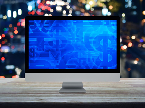 Financial currency symbol on desktop computer monitor screen on wooden table over blur colorful night light traffic jam road in city