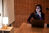 Young pretty smiling woman using laptop in home at night. Sad girl looking at computer. Cheerful people sitting at desk in living room. Concept of enterprising, telecommuting and hardworking person.