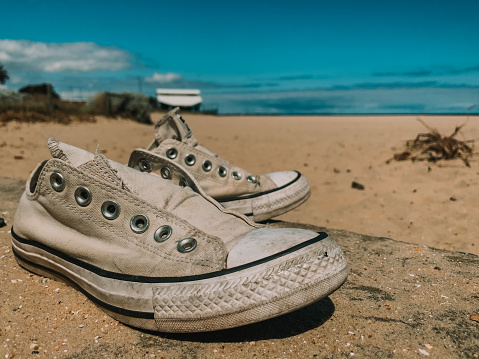 Old worn-out sneakers stand on the ocean shore, on a sandy beach, with a blue ocean in the background. Travel concept.