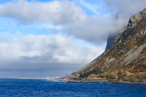 A looming mountain cliff extends into the ocean, partly shrouded by clouds under a blue sky. Cluster of houses by the water's edge