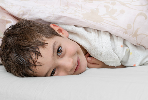 Smiling little boy hiding under blanket in bed after shower.