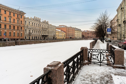 St. Petersburg, Russia, February 10, 2024. View of a river embankment in the city center, granite banks and a cast-iron fence with ornaments, historical buildings in a classical architectural style, black trees, cars and people walking, cityscape, seasonal view, snow and ice, winter.