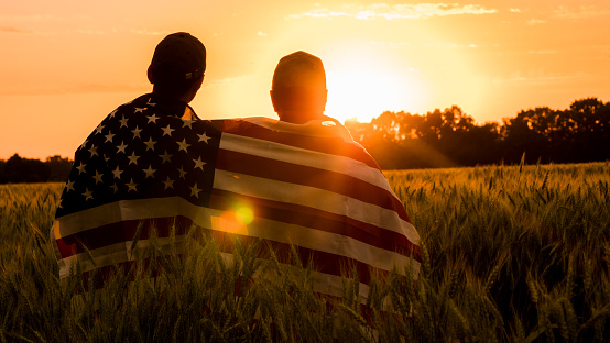 A man and his son admire the sunset over a field of wheat, wrapped in the flag of the USA.