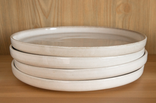 A stack of four beige ceramic dinner plates in a wooden cupboard.