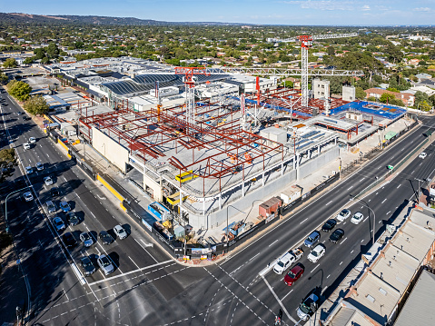 Aerial view large retail shopping mall construction site in Adelaide's eastern suburbs with two large tower cranes, construction equipment, building worksite, painted steel framework, concrete flooring, blue material.  Adjacent busy main road intersection with traffic buildup and leafy green suburbs and ocean in distance. ID and logos edited, photographed on a public holiday with no on-site activity.  (Burnside Village, corner Portrush & Greenhill Roads)