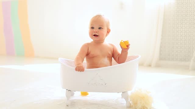 a little baby girl of six months with blue eyes bathes in a bubble bath and plays with rubber toys with ducks, smiles and rejoices splashing in the water, a happy baby washes