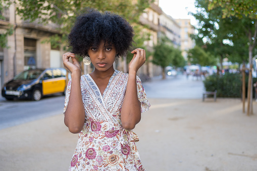 A young mixed-race woman with afro hair posing very shyly in front of the camera on the streets of Barcelona, Spain