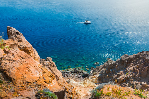Santorini island, Greece. Yacht sailing near the sea coast. Clear blue sea and red volcanic rocks. Summer seascape.