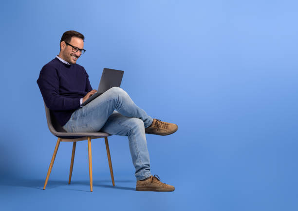 istock Confident young businessman in eyeglasses using laptop while sitting on chair over blue background 2125032609