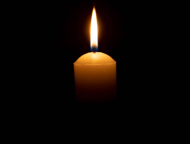 istock Single burning candle flame or light glowing on big white candle on black or dark background on table in church for Christmas, funeral or memorial service with copy space 2125132248