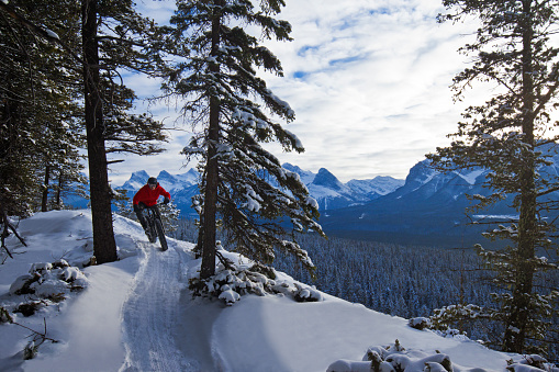 A man goes for a winter fatbike ride in the  Rocky Mountains of Canada. Fatbikes are mountain bikes with oversized wheels and tires for riding on the snow. He wears goggles, a bicycle helmet, and warm winter clothing. The Three Sisters mountain peaks, Ha Ling Peak and Mt Rundle are visible in the background.