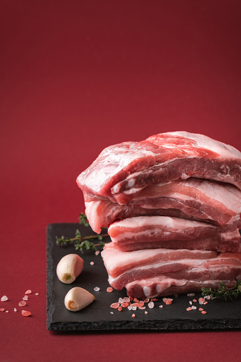 Close up shot of raw uncooked sliced pork meat stacked together surrounded by salt crystals, garlic and spices on the red background. Copy space for a free text. Cooking meat dishes recipe template.