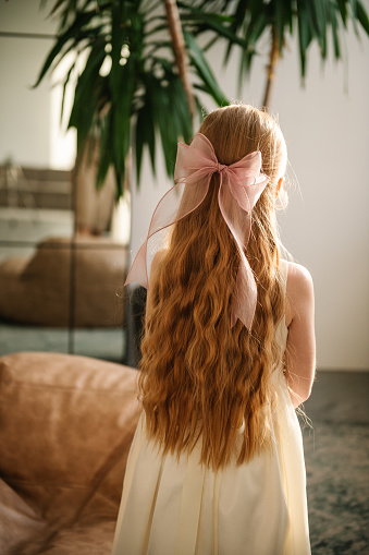 A beautiful girl with long curly red hair and a big bow on her head is standing back at home. Child in luxury dress in the room. Back view.
